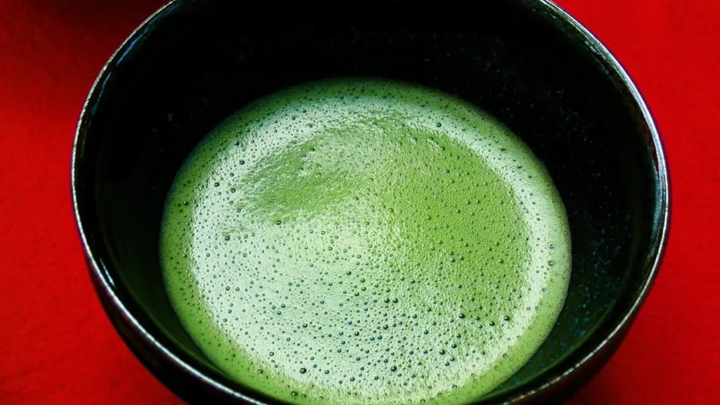 Matcha Tea Ratio Guide: Grams, Water and Whisking A ceramic Japanese chawan bowl filled with frothy bright green matcha tea beside a bamboo chasen whisk